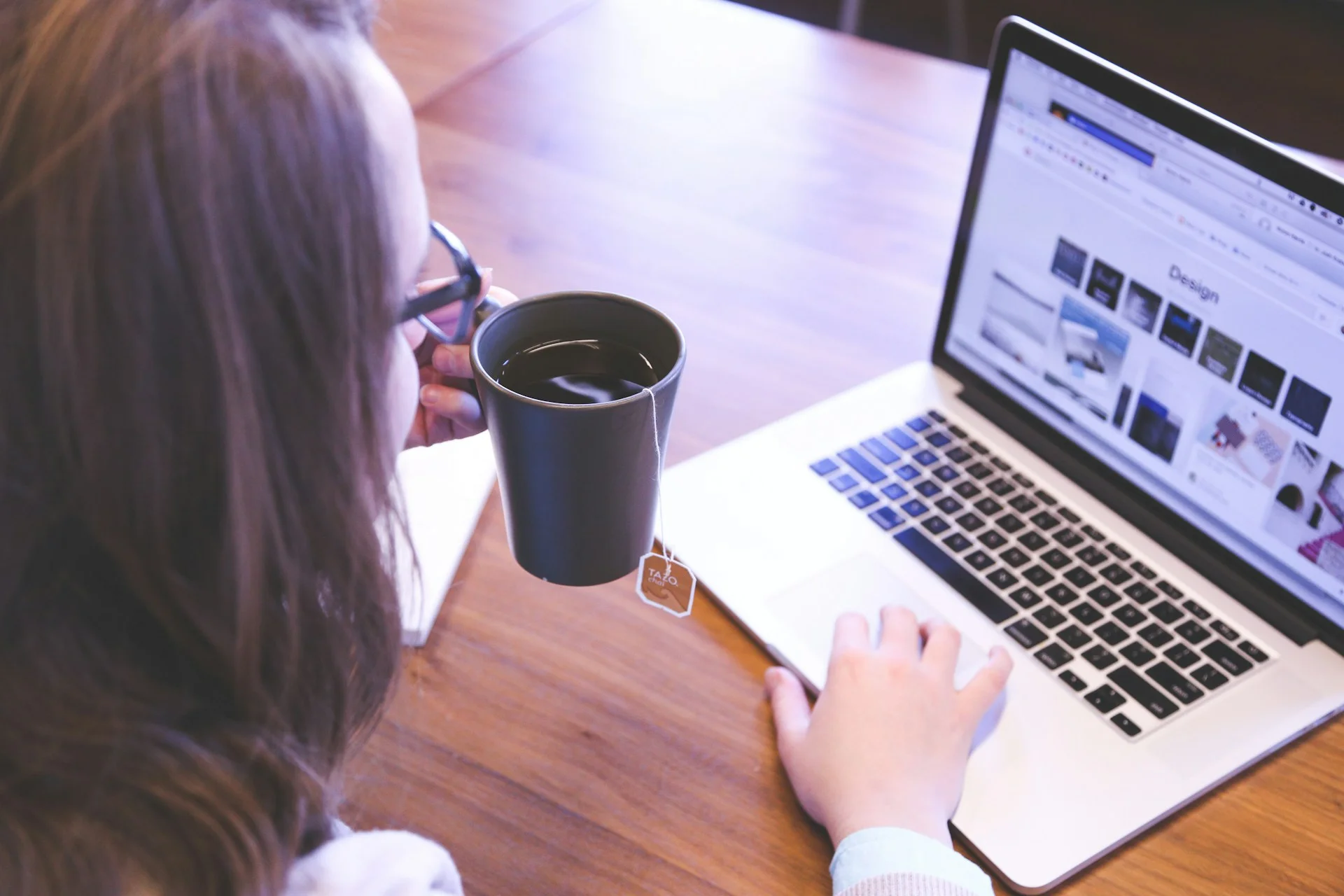Virtual assistant with glasses holding a mug while using a laptop on a wooden table.