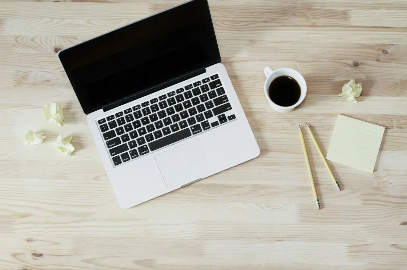 Virtual worker's laptop with a black screen, coffee cup, crumpled paper, and notepad on a wooden surface.